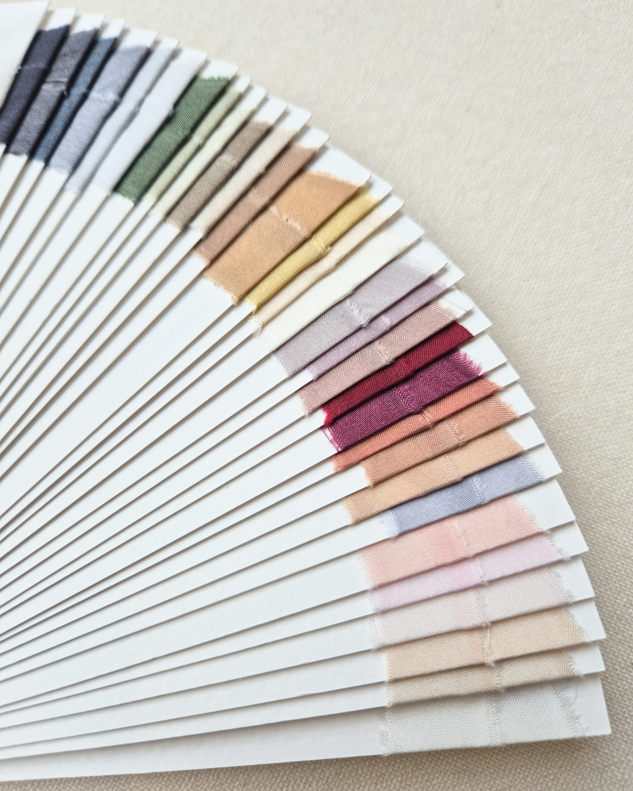 Close-up van handgeverfde zijden lintstalen in zachte neutrale tinten, roze, bordeaux, groen en blauw, met zichtbare stofstructuur.